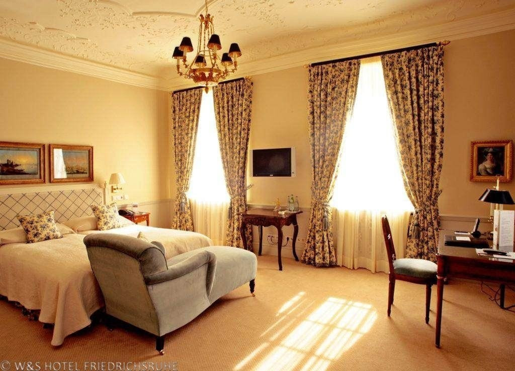 Blick in ein Hotelzimmer mit einem großen Bett auf der linken Seite und einer grauen Liegecouch davor. Die Decke ist mit Stuck verziert, auf der rechten Seite ist ein dunkler Schreibtisch aus Holz und ein Stuhl davor zu sehen.