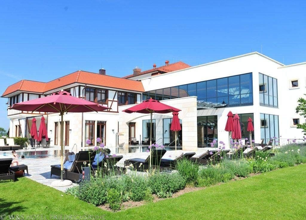 Ein Außenbereich eines Hotels mit Pool, davor stehen Ruheliegen am Pool und dunkelrote Sonnenschirme. IN der front ist ein Blumenbeet, im Hintergrund ist ein modernes Glasgebäude u sehen, das an einem ebenfalls modernen Gebäude in Fachwerkoptik grenzt.