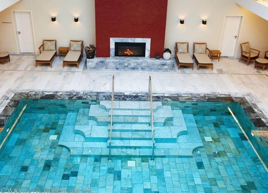 Blick von oben auf einen luxuriösen Indoor-Pool mit Steintreppe als Ein- und Ausgang für Schwimmer, Kamin vor dunkelroter Wand und Ruheliegen im Hintergrund.