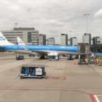 Ein KLM Cityhopper-Flugzeug steht auf dem Rollfeld des Flughafens von Amsterdam. In der Nähe stehen Gepäckwagen, im Hintergrund sind Terminalgebäude zu sehen.
