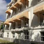 Das elegante weiße Gebäude mit dem Namen „Carlton Ambassador Hotel“ besticht durch klassische Architektur und gestreifte Markisen über den Fenstern. Es liegt in Den Haag und steht auf einer Rasenfläche, in deren Nähe sich ein Laternenpfahl befindet.