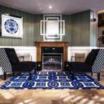 In einem gut beleuchteten Raum stehen zwei schwarz gemusterte Sessel vor einem Kamin auf einem blauen geometrischen Teppich, flankiert von einem eleganten Kaminsims aus Holz und geschmückt mit Kunstwerken an den Wänden.