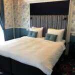 In einem gemütlichen Hotelzimmer in Den Haag steht ein ordentlich gemachtes Doppelbett vor einer gestreiften und geblümten Tapete, mit blau-weißer Bettwäsche und zwei blaugrünen Kissen.