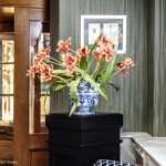 Eine blau-weiße Porzellanvase mit rosa Blumen thront elegant auf einem schwarzen Sockel vor einer grün gestreiften Wand, die mit gerahmten Kunstwerken und dekorativen Tellern geschmückt ist und an ein schickes Den Haag-Interieur erinnert.