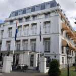 Ein weißes mehrstöckiges Gebäude in Den Haag verfügt über gestreifte Markisen und Fahnenmasten am Eingang sowie Sitzbereiche im Freien, umgeben von üppigem Grün.