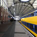 Ein gelb-blauer Zug steht unter einem gewölbten Bahnsteig mit Glasdach in Den Haag. In der Nähe warten einige Passagiere.