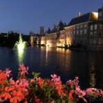 Ein Blick auf einen Kanal in Den Haag in der Abenddämmerung, mit beleuchteten Gebäuden, die sich im Wasser spiegeln, leuchtenden Blumen im Vordergrund und einem beleuchteten Brunnen, der in der Ferne sanft leuchtet.