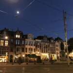 Die Nacht bricht über einen Stadtplatz in Den Haag herein, über dem sich beleuchtete Straßenbahnleitungen winden, historische Gebäude stolz aufragen und eine Statue wacht. Mond und Straßenlaternen werfen gemeinsam einen ruhigen Schein über die Gegend.