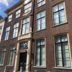 Dieses historische Gebäude aus braunem Backstein liegt im Herzen von Den Haag und verfügt über hohe Fenster, einen dekorativen Eingang und eine kleine Treppe mit Geländer, die Sie zur schwarzen Tür führt.