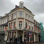 Ein Eckcafé in Den Haag im Erdgeschoss eines dreistöckigen Backsteingebäudes bei bewölkten Himmel. Menschen stehen draußen, während Motorroller in der Nähe geparkt sind und zur lebhaften Szene beitragen.