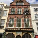 Historisches niederländisches Gebäude in Den Haag mit dekorativer Backsteinfassade, großen Fenstern und einem Stufengiebel. Unter dem sonnigen blauen Himmel mit Wolken sind eine Straßenlaterne und Leitungen zu sehen.