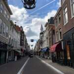 Eine schmale, leere Straße in Den Haag ist gesäumt von Backsteingebäuden unter einem teilweise bewölkten Himmel, geschmückt mit bezaubernden Hängelaternen.