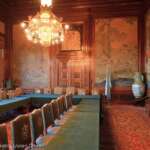 Eleganter Raum in Den Haag mit langen, mit grünem Tuch bedeckten Tischen. Holzstühle stehen auf beiden Seiten, während ein verzierter Kronleuchter von der Decke hängt. Die Wände sind mit großen Wandgemälden geschmückt, und eine dekorative Vase ziert die Ecke, was diesem charmanten Raum einen Hauch von Raffinesse verleiht.