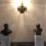 Zwei Büsten auf Sockeln unter einem dekorativen Wandleuchter in Den Haag: eine mit der Aufschrift „Nelson Mandela“ und die andere trägt den Namen „Mahatma Gandhi“, aufgestellt in einem hell erleuchteten Raum mit kunstvollen Details.