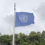 Die Flagge der Vereinten Nationen weht anmutig auf einem Mast vor einem bewölkten Himmel und einer Baumreihe und erinnert an den Geist der internationalen Diplomatie, wie er in Den Haag herrscht.