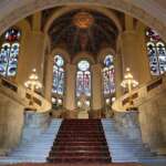 Die große Marmortreppe des Friedenspalastes in Den Haag mit ihrem roten Teppich und den kunstvollen Buntglasfenstern führt unter Kronleuchtern hindurch zu einer großen Kuppel, die mit Mustern geschmückt ist.