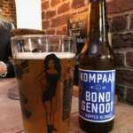 Ein Glas bernsteinfarbenes Bier steht neben einer Flasche Kompaan Bondgenoot Hopped Blond-Bier auf einem Holztisch in Den Haag, im Hintergrund sind eine Backsteinwand und eine Menütafel zu sehen.