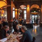 Menschen sitzen an Tischen in einem schwach beleuchteten Restaurant mit Backsteinwänden und gewölbten Türen in Den Haag, unterhalten sich und genießen Getränke.