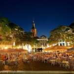 Die Nacht bricht über den Grote Markt in Den Haag herein, wo im Freien Tische, Stühle und Sonnenschirme sanft von Lichterketten beleuchtet werden. Im Hintergrund wacht ein Kirchturm über die geschäftige Szene.
