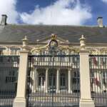 Großes weißes neoklassizistisches Gebäude mit verzierten Toren und einem Wappen mit einem blauen Löwen und einer goldenen Krone, das an die architektonische Eleganz Den Haags erinnert. Das Dach ist grau und ragt majestätisch unter dem teilweise bewölkten Himmel empor.