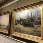 In einer Galerie in Den Haag hängen zwei goldgerahmte Gemälde mit Booten nebeneinander; eines fängt eine Hafenszene mit sanftem Licht ein, während das andere angedockte Boote unter einem bewölkten Himmel zeigt. Beide Kunstwerke werden elegant auf einer grünen Wand präsentiert.