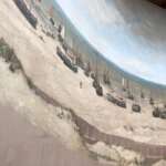 Ein Panorama-Wandgemälde fängt eine wunderschöne, historische Strandszene in Den Haag ein. Es zeigt zahlreiche Boote, die auf dem Sand liegen, während sich unter einem bewölkten Himmel das Meer bis zum Horizont erstreckt.