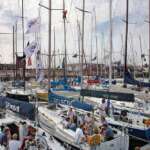 Im Yachthafen von Den Haag liegen viele Segelboote. Menschen sind an Bord und unterhalten sich. Flaggen flattern vor einem bewölkten Himmel.