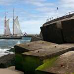 Ein Segelboot mit niederländischer Flagge gleitet durch das Wasser neben großen Betonblöcken am Ufer in Den Haag. Zwei Menschen schlendern einen betonierten Weg entlang, der sich oberhalb des Strandes windet.