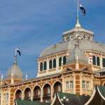 Das historische Gebäude von Den Haag verfügt über eine große Kuppel und kleinere Türme, deren Flaggen stolz vor einem klaren blauen Himmel wehen, während im Hintergrund eine moderne Struktur mit einer goldenen Kugel schimmert.