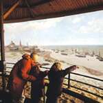 Eine Frau steht mit ihren beiden Kindern auf einer Besuchergalerie. Gemeinsam bewundern sie ein großes Panoramagemälde einer Strandszene mit Booten und Gebäuden, das an den historischen Küstencharme von Den Haag erinnert.