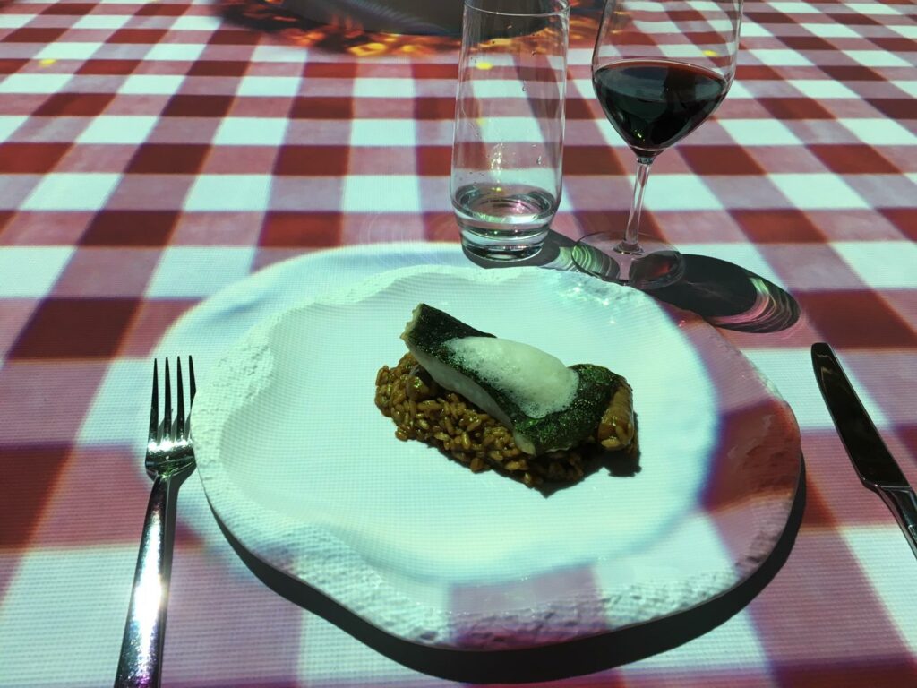 Ein Gericht von TastEmotion präsentiert ein Stück Fisch in weißer Soße auf gewürzten Körnern auf einem weißen Teller. Gabel und Messer liegen ordentlich daneben, begleitet von einem Glas Wasser und Rotwein auf einer rot-weiß karierten Tischdecke, was die perfekte Stimmung für ein exquisites Speiseerlebnis schafft.