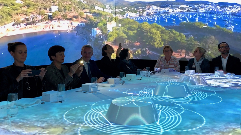 Menschen sitzen in einem Raum um einen Tisch herum, auf den Bilder einer Küstenszene projiziert werden.