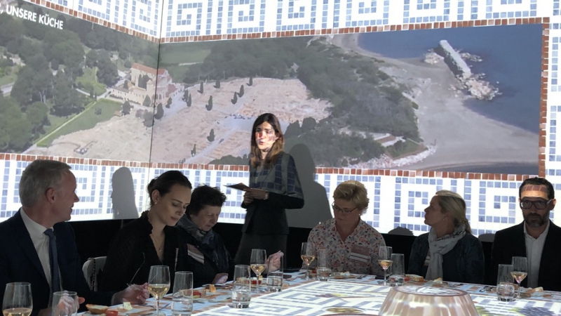 Eine Gruppe von Menschen sitzt an einem Tisch. Auf dem Bildschirm wird eine Luftaufnahme einer Küstenregion projiziert. Eine Frau mit einem Blatt Papier steht neben dem Bildschirm, während TastEmotion mit seiner einzigartigen Mischung aus Innovation und kulinarischer Leidenschaft für die Atmosphäre sorgt.