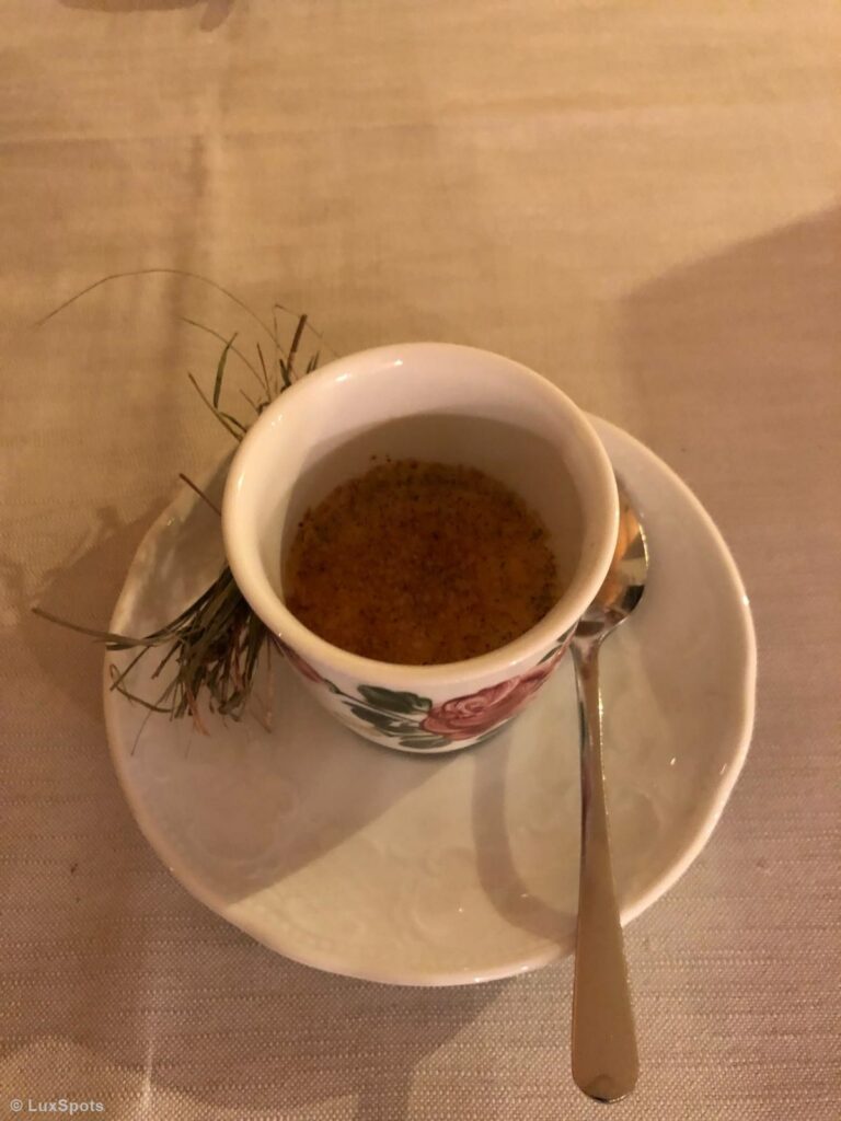 Eine weiße Espressotasse mit Crème Brûlée auf einer Untertasse mit Löffel, als Deko liegt etwas grünes Heu auf einer beigen Tischdecke.