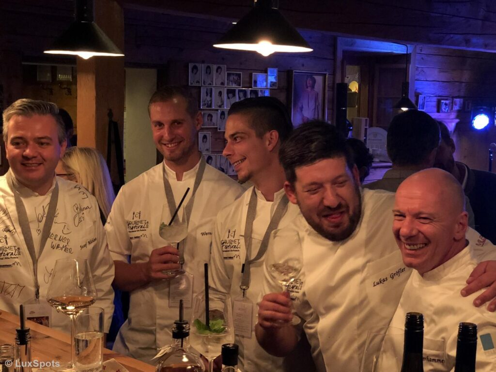Eine Gruppe von Köchen in weißen Uniformen sitzt bei einer schwach beleuchteten Veranstaltung zusammen, lächelt und hält Getränke in den Händen. Flaschen und Gläser stehen auf dem Tisch vor ihnen.