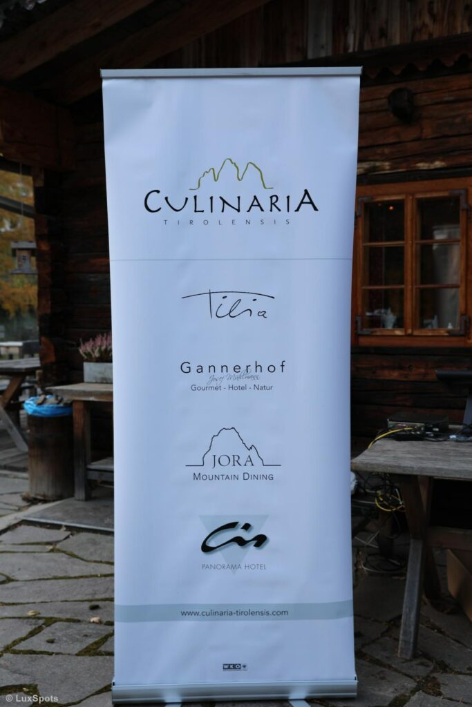 Ein Banner mit den Logos von Culinaria Tirolensis, Tilia, Gannerhof, Jora Mountain Dining und Panorama Hotel vor einem rustikalen Holzgebäude.