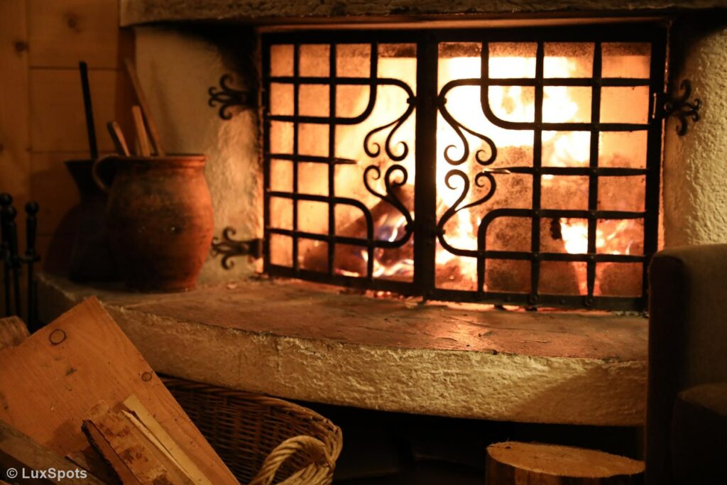 Ein brennender Kamin mit schmiedeeisernem Gitter, umgeben von Holzscheiten und einem Korb, schafft eine gemütliche Atmosphäre.