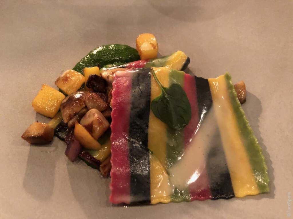 Bunt gestreifter Raviolo mit heller Sauce, garniert mit einem Blatt, umgeben von Gemüsewürfeln und Pilzen auf einem beigen Teller.