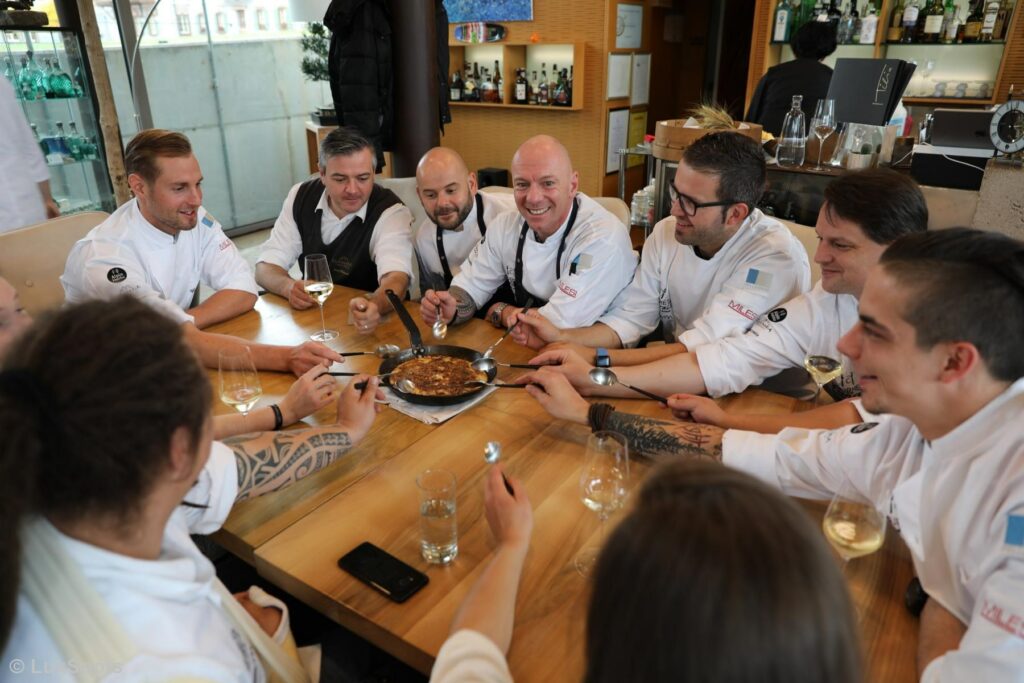 Eine Gruppe von Köchen in weißen Uniformen teilt sich begeistert ein südtiroler Gericht an einem Holztisch in einem Restaurant.