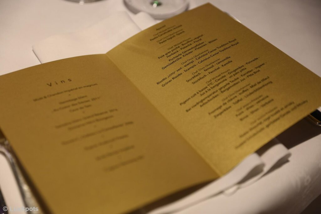 Auf einem Tisch liegt eine aufgeschlagene Speisekarte, auf der auf goldfarbenem Papier gedruckt verschiedene Weine und Speisen des Abends aufgeführt sind.