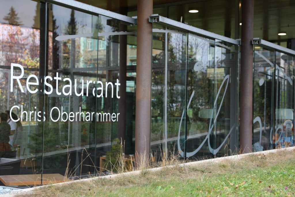 Außenansicht eines Restaurants im Hochpustertal mit großen Fenstern, auf denen elegant der Name „Chris Oberhammer“ prangt. Grünes Gras und üppige Bäume umgeben das moderne Lokal.
