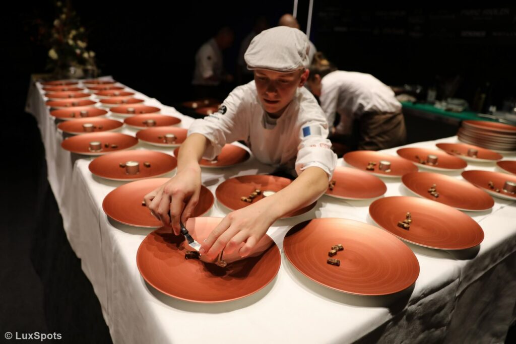 Ein Koch in weißer Uniform und Mütze arrangiert sorgfältig Speisen auf orangefarbenen Tellern, die auf einem großen Tisch verteilt sind.