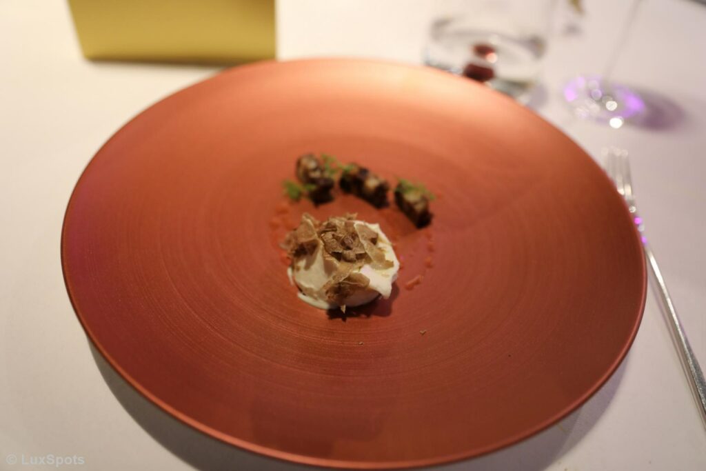 Ein roter Teller mit einem Gourmetgericht aus Trüffeln auf cremiger weißer Soße und kleinen garnierten Süigkeiten, auf einer weißen Tischdecke gedeckt, mit einer Gabel und einem Glas Wasser daneben.