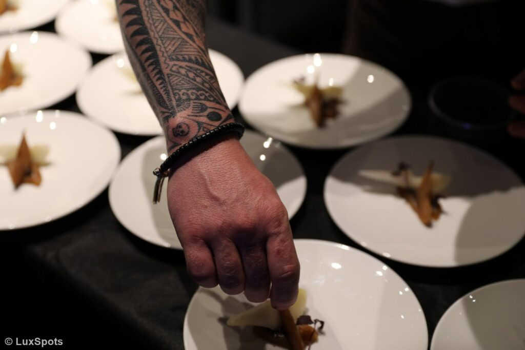 Eine tätowierte Hand arrangiert kunstvoll Speisen auf weißen Tellern, auf denen sich jeweils ein kleines, feines Schälchen befindet.