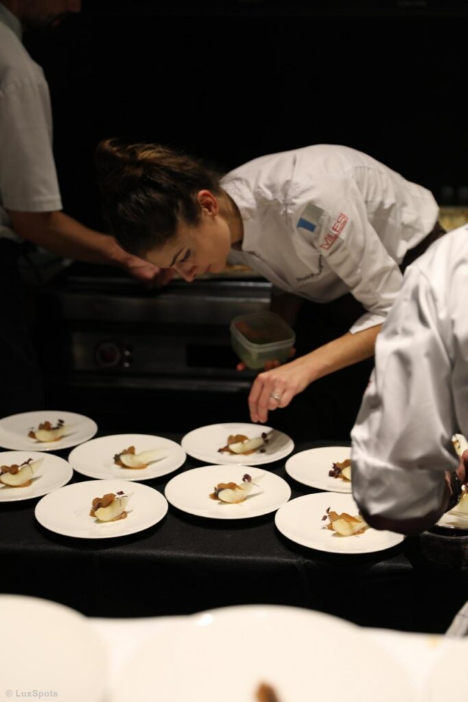 In einer schwach beleuchteten Küche arrangiert eine Patissière sorgfältig Desserts auf mehreren Tellern.