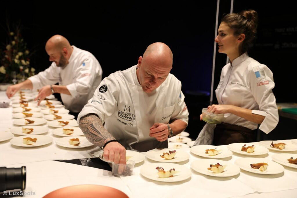 Bei einem kulinarischen Event richten Köche die Desserts sorgfältig an und arrangieren die Elemente in einem gut beleuchteten Ambiente auf mehreren Tellern.