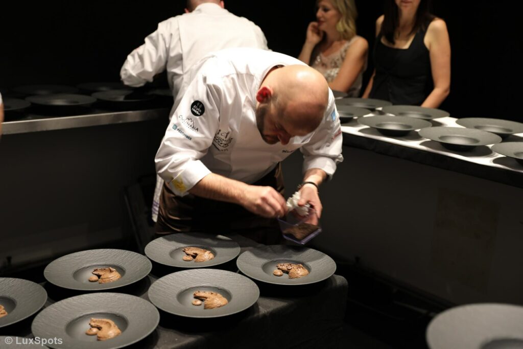 Ein Koch im weißen Kittel garniert in einer schwach beleuchteten Küche sorgfältig Dessertteller, im Hintergrund sind mehrere Personen zu sehen.