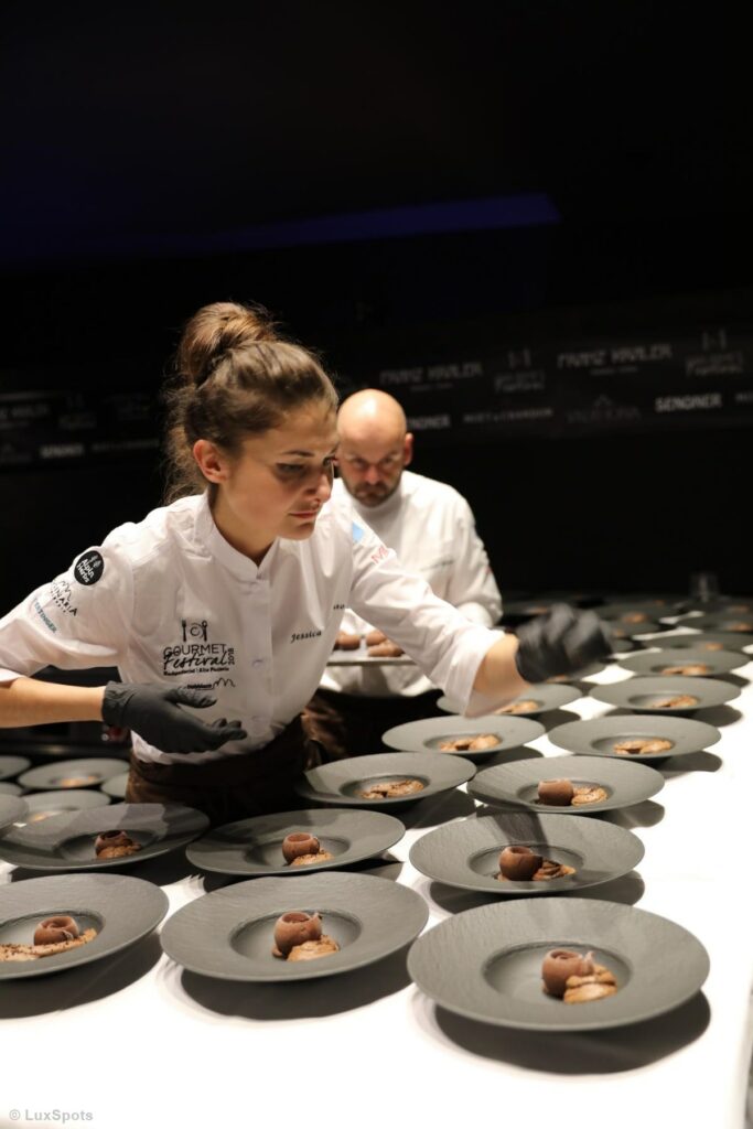 Eine Patissière richtet sorgfältig mehrere Teller mit Schokoladendesserts, während ein Koch im Hintergrund arbeitet.