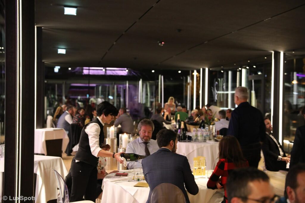 In einem belebten, modernen Restaurant mit gedämpfter Beleuchtung und vielen Gästen schenkt ein Kellner einem sitzenden Gast Wein ein.