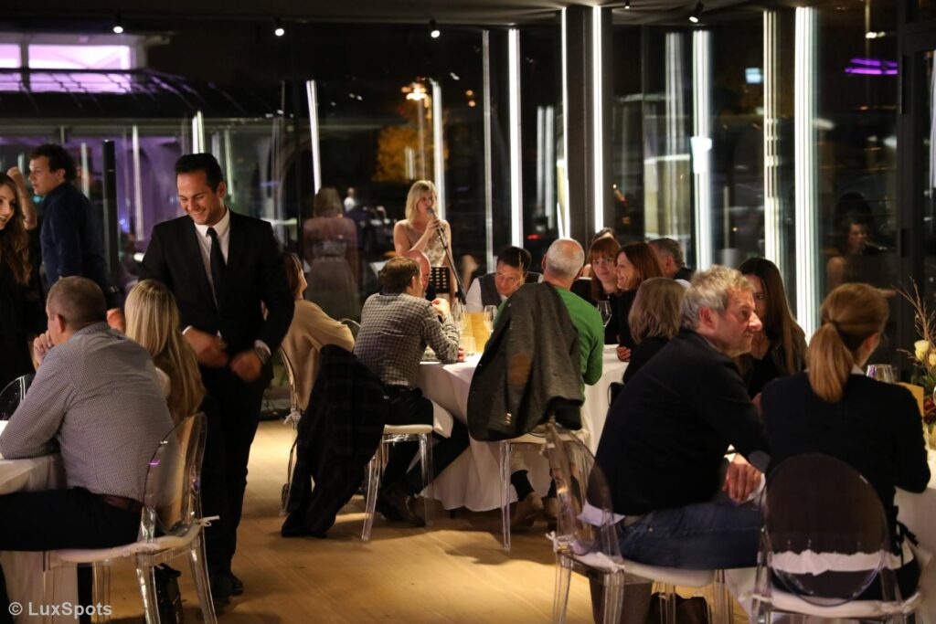 Eine lebhafte Restaurantszene mit Menschen, die an Tischen vor großen Fenstern im Hintergrund speisen. Im Hintergrund ist eine Sängerin zu sehen.